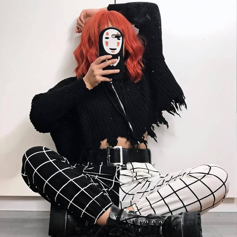 Pantalon gothique femme noir et blanc DarkWhiteWave avec motif à carreaux, moitié noir à carreaux blancs et moitié blanc à carreaux noirs, porté assis avec ceinture noire et bottes noires