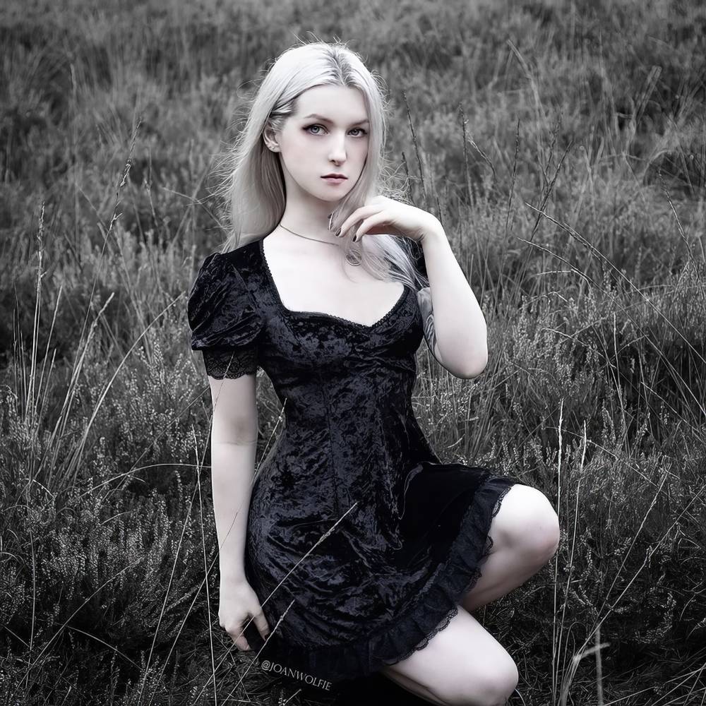 Femme portant une robe gothique chic noire en velours avec manches bouffantes et décolleté carré, modèle CyberClash, assise dans l'herbe