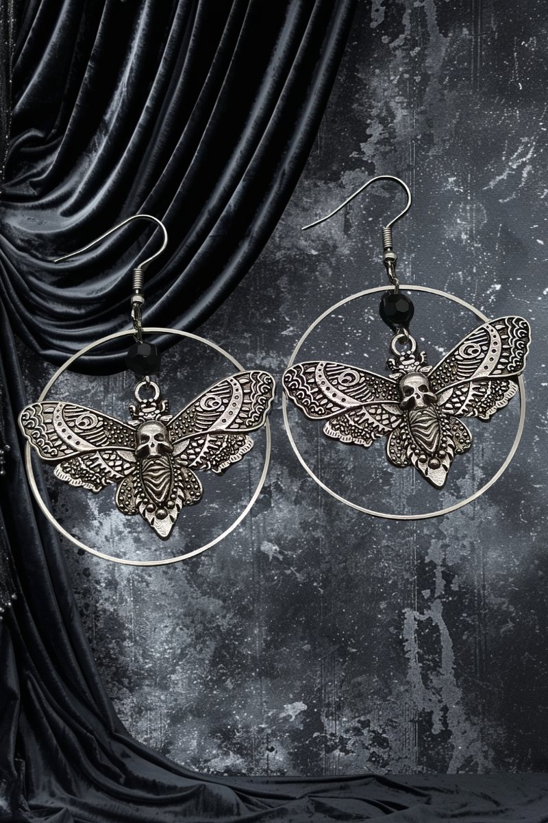 Boucles d'oreilles gothiques Phantomglow en métal argenté avec motif papillon et tête de mort, suspendues sur anneaux circulaires, fond noir texturé avec tissu velours noir