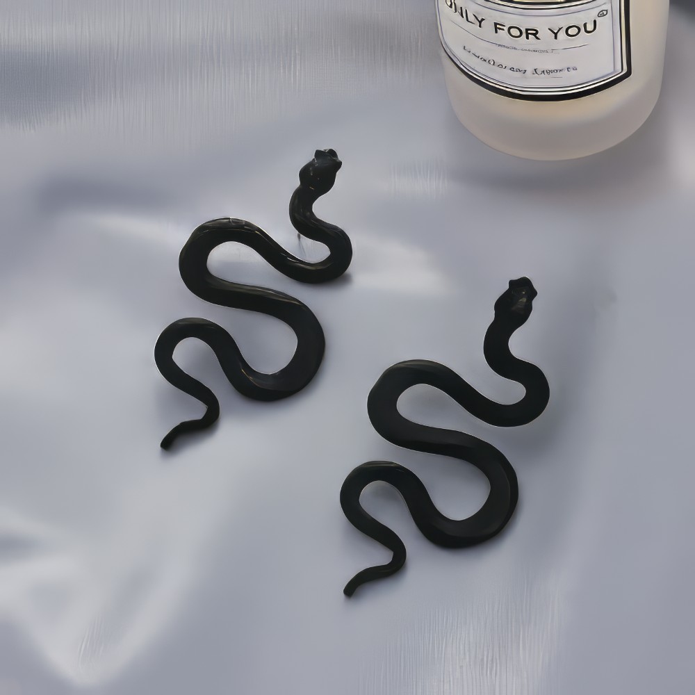 Boucles d'oreilles gothiques serpent en S noires en métal mat modèle Ophidianeclipse posées sur tissu blanc avec bougie en arrière-plan